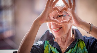Older woman in blue shirt and green scarf making a heart with her fingers around her eye